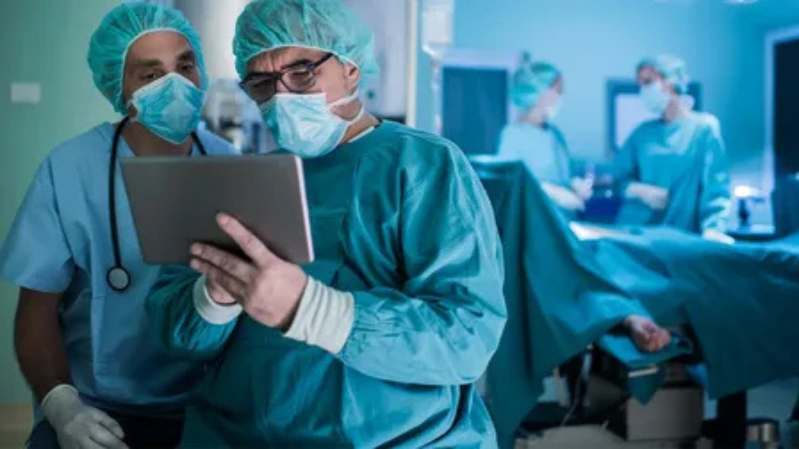 Two doctors in surgical attire, including masks and scrubs, are examining a tablet in a medical operating room.